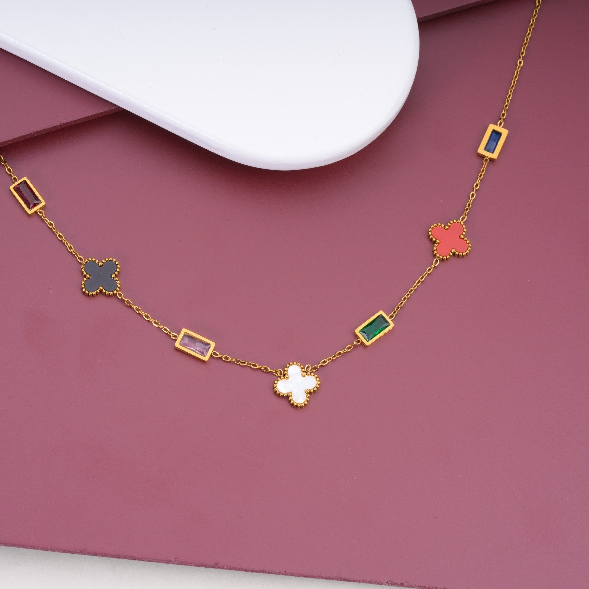 Elegant gold chain necklace with multicolored rectangular and clover-shaped charms on mauve background