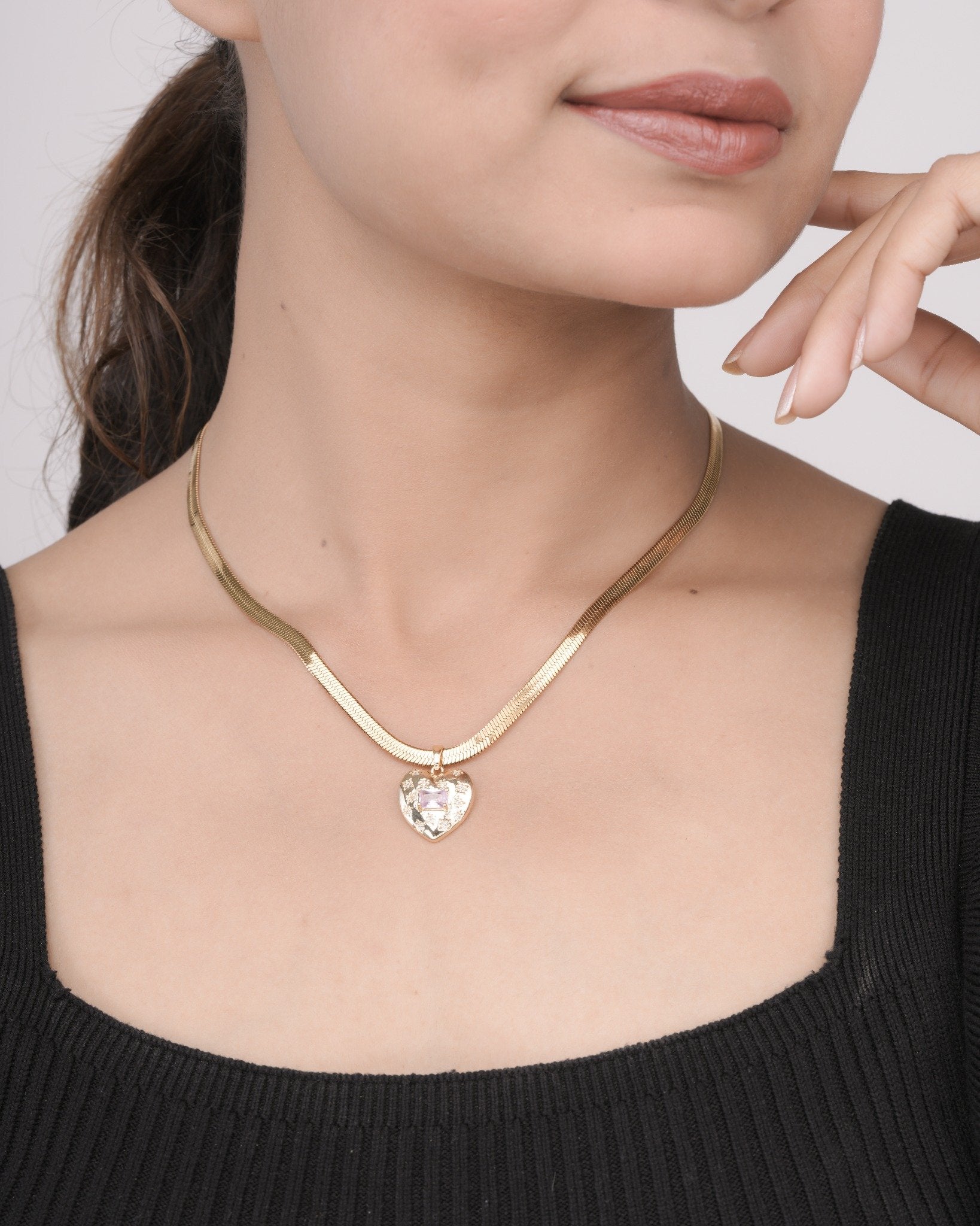 Close-up of woman wearing a gold heart-shaped pendant necklace with a pink gem on a gold chain, paired with black top