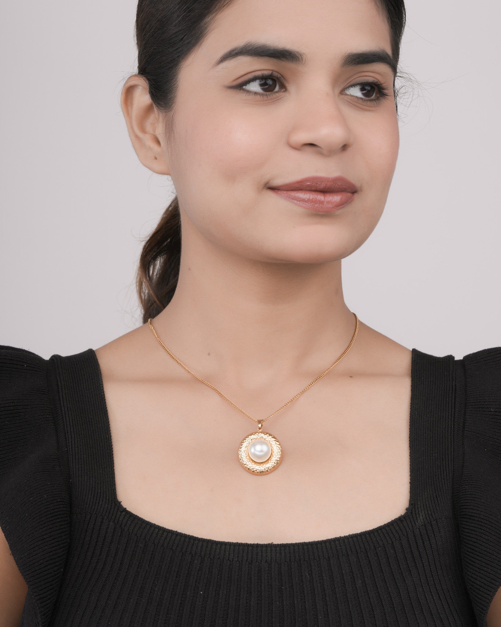 Woman wearing a gold chain necklace with a round pendant featuring a central white pearl on a black top