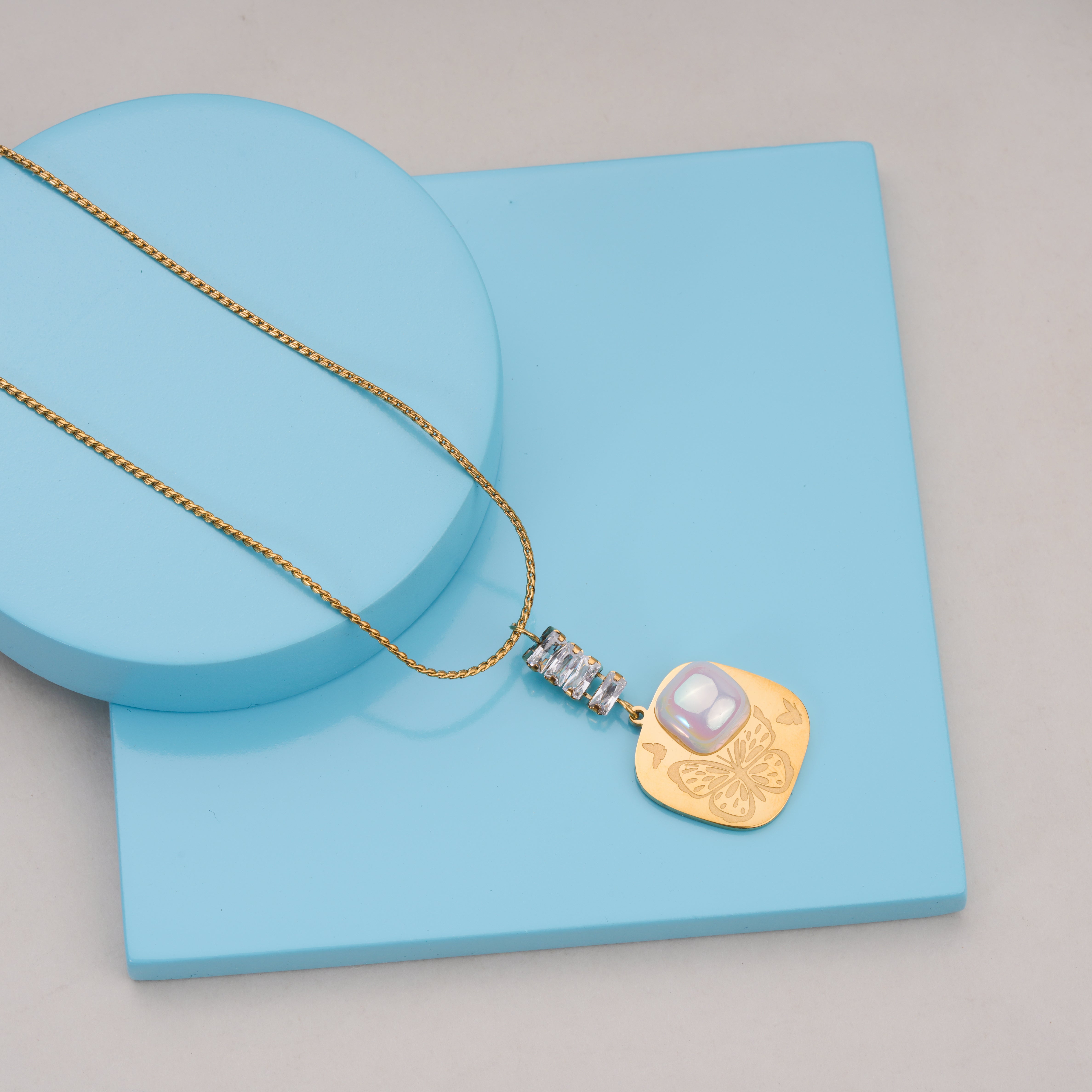 Gold chain necklace with pendant featuring butterfly engraving, pearl detail, and crystal accents on blue display