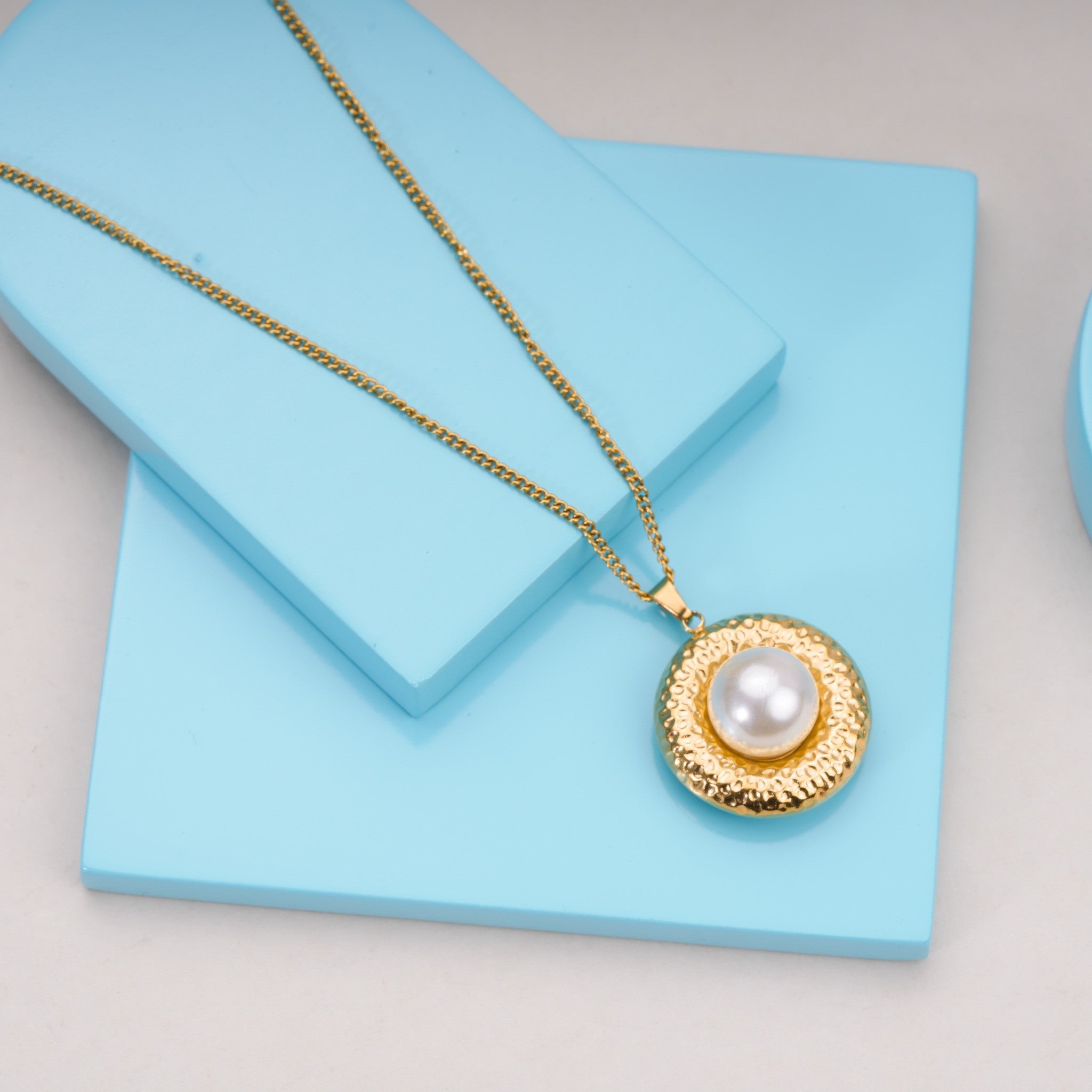 Gold pendant necklace with large white pearl on textured round charm, displayed on pastel blue blocks