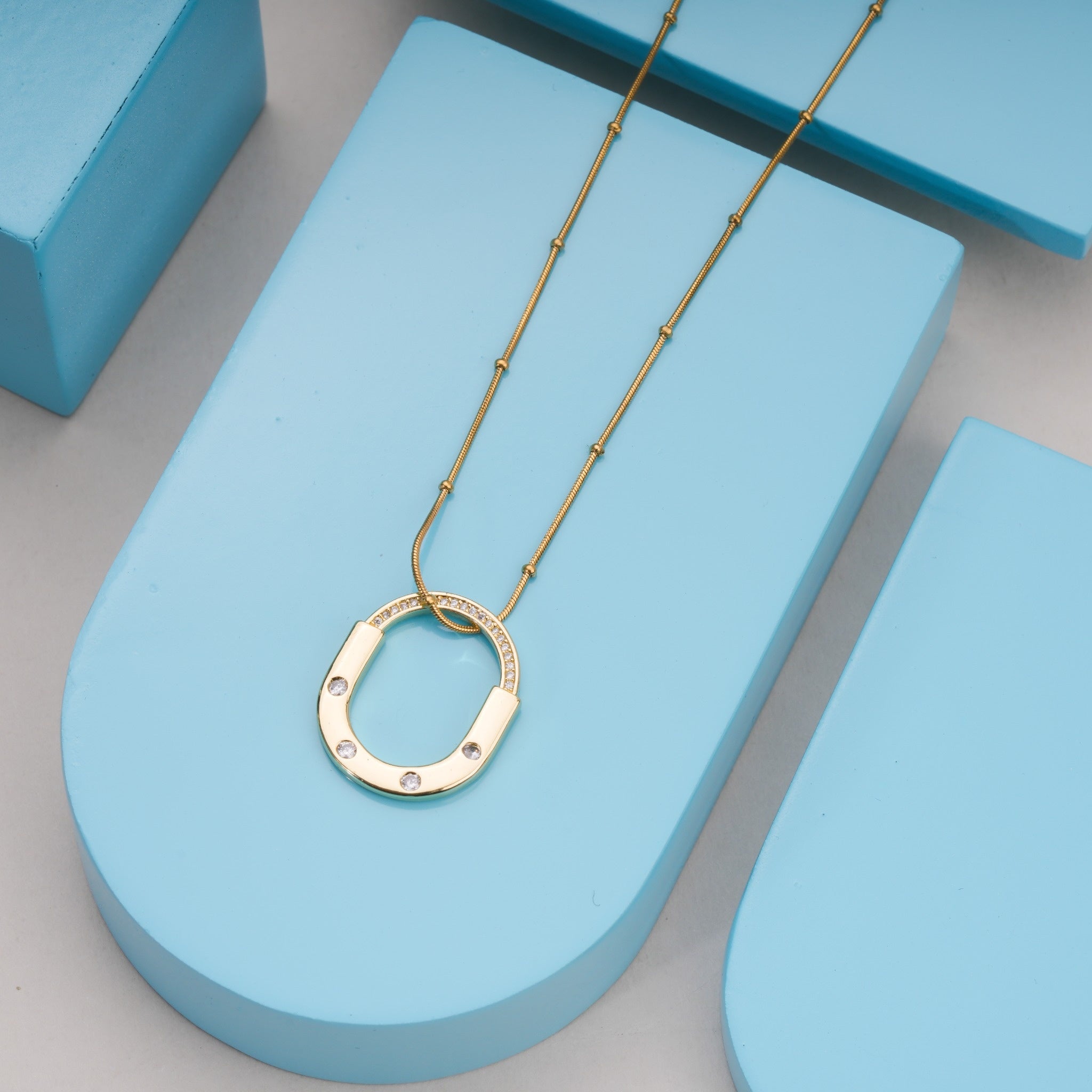 Gold chain necklace with oval pendant featuring embedded crystals on blue geometric display
